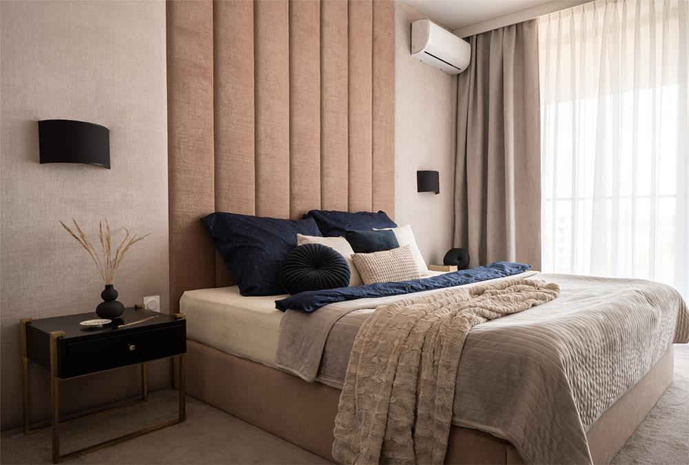 Bedroom with a beige upholstered wall and bed, a black bedside table, and a black wall lamp. Air conditioning in the corner of the wall.