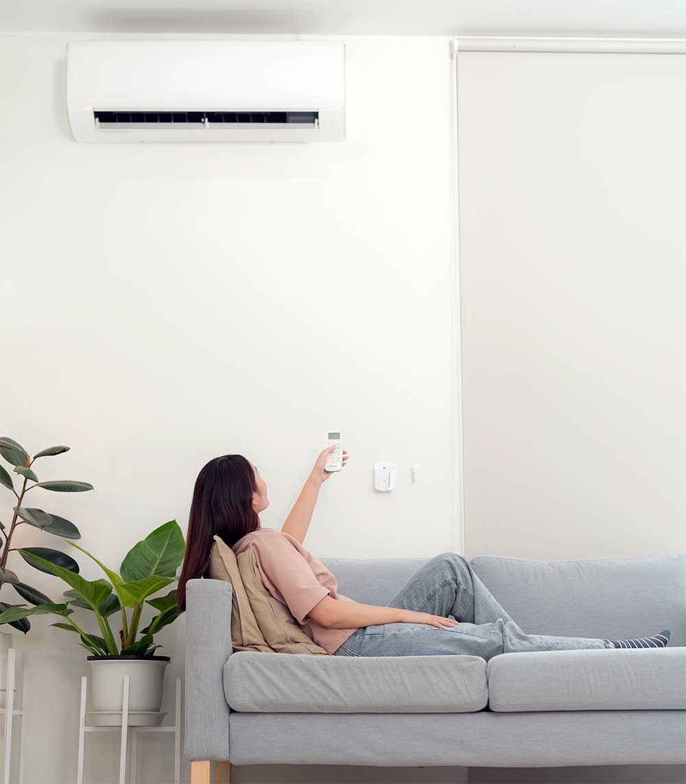 Woman on a couch using a remote control to adjust an air conditioner mounted on the wall.